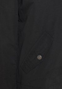 Sort jakke med en tekstureret overflade, der har en sidelomme med en metallisk knapdetalje, samt et minimalistisk og funktionelt design.