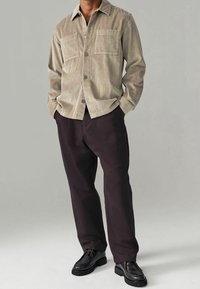 Chemise boutonnée en velours côtelé beige avec deux poches avant et pantalon marron foncé. La chemise présente un motif texturé et un col. Chaussures noires.