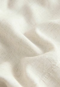 Makro záber mäkkej, prírodnej béžovej látky s viditeľnou tkanou textúrou a jemnými záhybmi vytvárajúcimi jemné tiene.