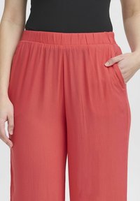 Rote, locker sitzende Hose aus strukturiertem Stoff, mit einem elastischen Bund und seitlichen Taschen, gezeigt mit einer entspannt ruhenden Hand.