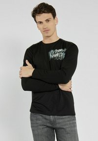 Czarna koszula z długim rękawem i okrągłym dekoltem, z nadrukowanym graficznym wzorem w białym i niebieskim kolorze, noszona z szarymi dżinsami.