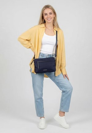 Junge Frau trägt gelbes Hemd, weißes Oberteil, hellblaue Jeans, weiße Sneakers und eine marineblaue Umhängetasche, lächelt und steht auf einem Bein.