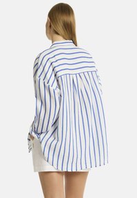 Una persona con capelli lunghi indossa una camicia a righe larga con linee blu e bianche, rivelando uno stile casual, abbinata a pantaloni corti bianchi.