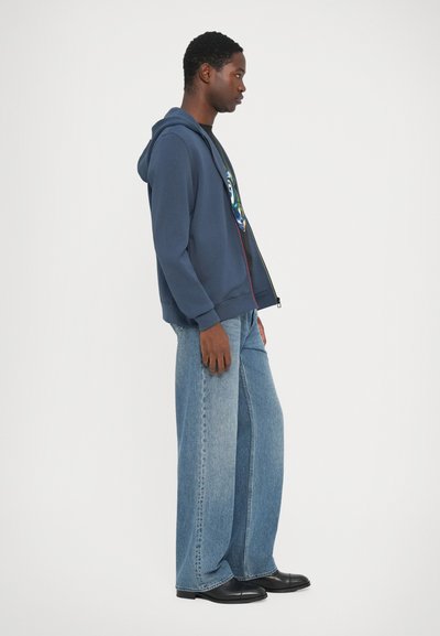 Felpa con zip di colore navy, dotata di tasche laterali, abbinata a jeans leggeri a gamba larga di colore blu chiaro e scarpe nere. Il tessuto sembra morbido, dal design casual.
