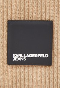 Pachet de țesătură rectangulară neagră cu text alb în relief care citește "KARL LAGERFELD JEANS." Fundalul prezintă o țesătură bej texturată.