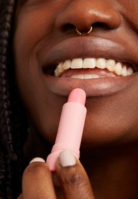 En rosa leppestift tube holdt nær leppene, med en avrundet spiss. Glatt tekstur og minimalistisk design, med en kremfarget hånd som holder den.