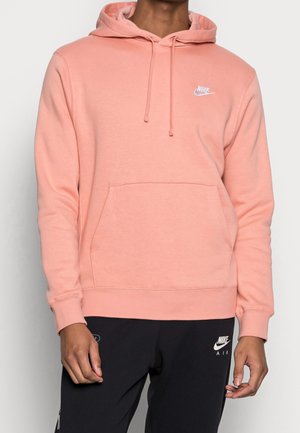 Osoba ubrana w jasnoróżową bluzę Nike z kapturem i kieszenią z przodu oraz czarne spodnie dresowe Nike Air.