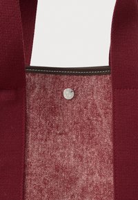 Τσάντα χειρός σε βαθυκόκκινο με υφή υφάσματος, αντίθετη ραφή σε χρώμα ταμπά και ασημένιο κουμπί σκρατς στο πάνω μέρος.