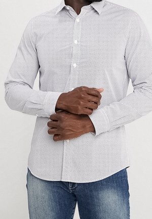 Mann trägt ein weißes gemustertes Hemd mit Knöpfen und eine blaue Jeans und richtet mit der rechten Hand die Manschette des linken Ärmels.