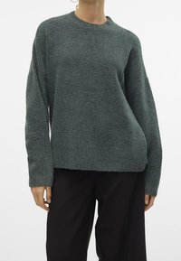 Nainen, jolla on yllään tummanvihreä neulepaita pitkillä hihoilla ja mustat housut, taustanaan valkoinen yksinkertainen tausta.