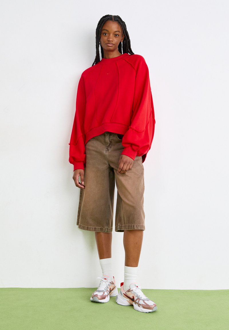 Felpa rossa oversize con polsini a costine, abbinata a pantaloni corti marroni che arrivano al ginocchio e sneaker con dettagli argentati su un superficie verde.