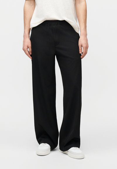Personne portant un pantalon noir ample avec une taille à cordon et des baskets blanches, associé à un t-shirt blanc rentré.