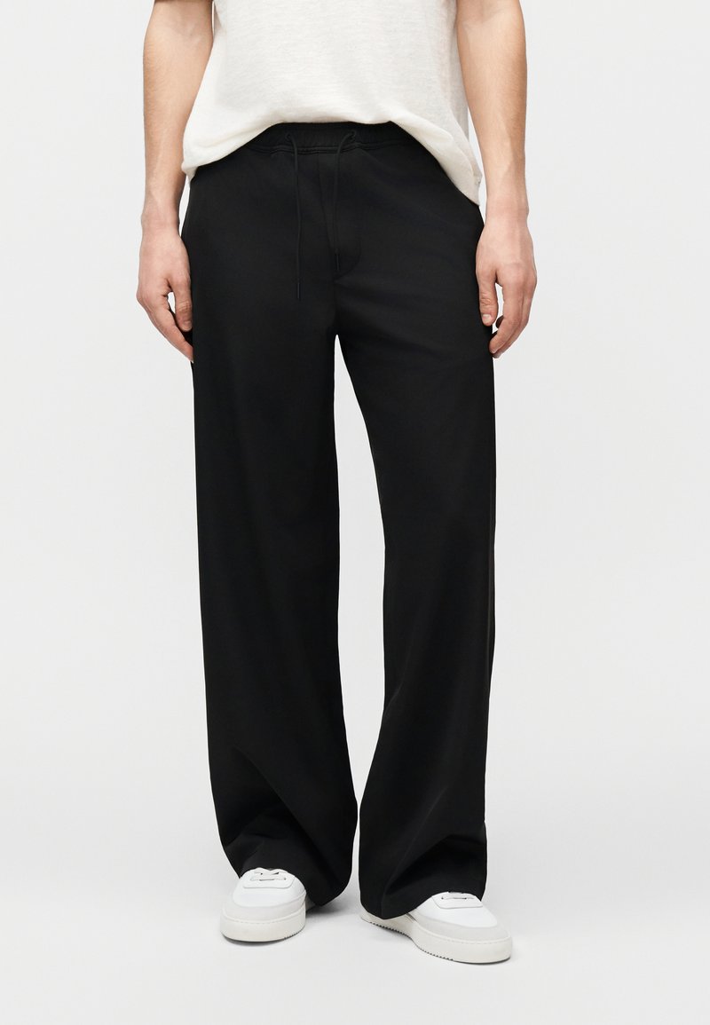 Άτομο που φοράει φαρδιά μαύρη παντελόνια με κορδόνι στη μέση και λευκά αθλητικά παπούτσια, συνδυασμένα με λευκό μπλουζάκι φορεμένο μέσα από το παντελόνι.