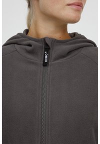 Chaqueta de forro polar gris oscuro con cierre frontal, capucha y etiqueta de marca en el cuello. La textura del tejido parece suave y cálida.