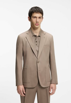 Giovane uomo che indossa un abito beige con tasche applicate e una polo abbottonata abbinata, in piedi davanti a uno sfondo bianco uniforme.