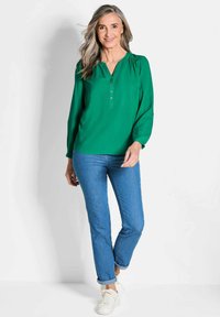 Blusa verde a maniche lunghe con scollo a V e bottoni, abbinata a jeans blu e sneakers bianche. Il tessuto sembra liscio; la blusa presenta una leggera arricciatura alle spalle.