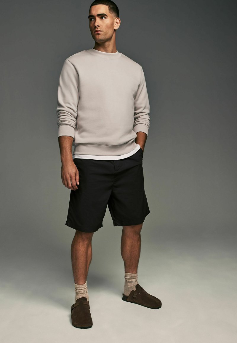 Sudadera gris claro, pantalones cortos negros, calcetines beige y zapatos marrones sin cordones. Diseño simple sin patrones, enfocado en la comodidad y el estilo casual.