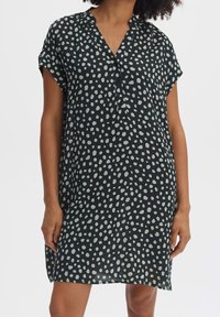 Femme portant une robe noire ample avec des pois blancs irréguliers, des manches courtes et un col en V avec des boutons sur un fond uni.