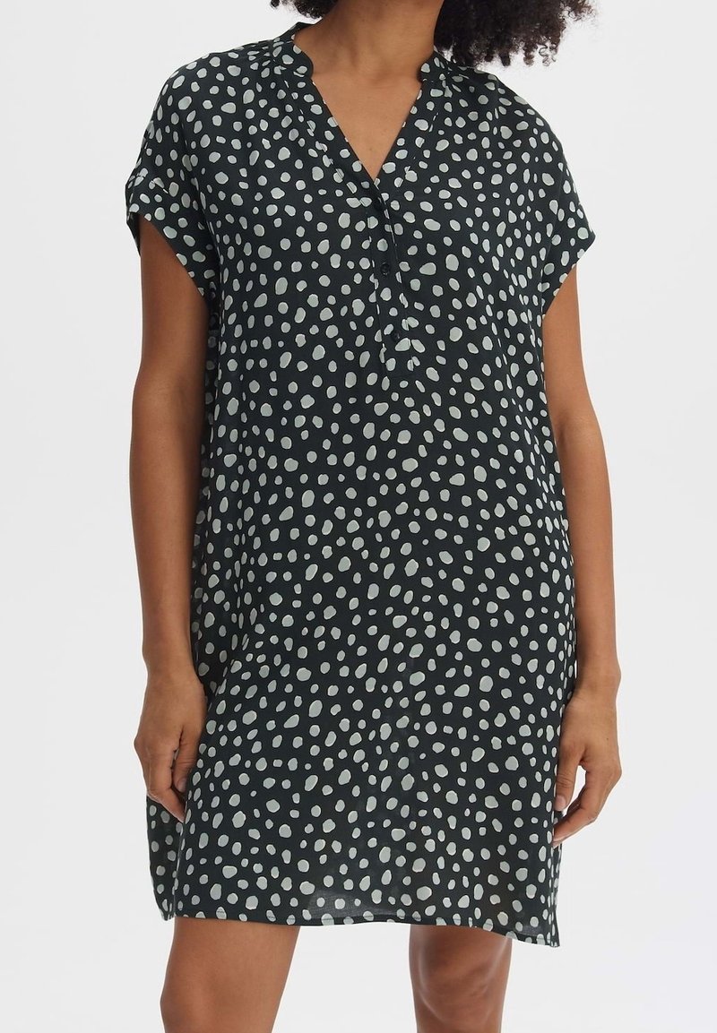 Femme portant une robe noire ample avec des pois blancs irréguliers, des manches courtes et un col en V avec des boutons sur un fond uni.