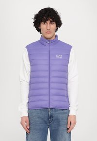 Colete puffer de cor lilás claro com gola alta, fecho de correr completo e logotipo EA7; texturizado com costuras horizontais; usado sobre uma camisa de manga longa branca.