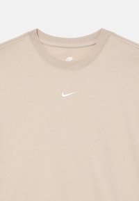 Beżowa koszulka Nike z okrągłym dekoltem, z białym haftowanym logo Nike swoosh umieszczonym na środku poniżej dekoltu.
