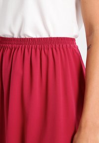Taille d'une personne portant un haut blanc rentré dans une jupe taille haute à taille élastique de couleur rose profond, avec un bras droit tatoué visible.