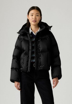 Jonge vrouw in een zwarte pufferjack over een gebreide trui en een wit shirt, staand tegen een effen lichtgrijze achtergrond.