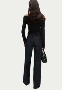 Wijdvallende donkere spijkerbroek met twee achterzakken en gouden stiksels, gecombineerd met een off-shoulder zwarte top en een kleine zwarte handtas.