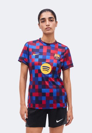 Person iført rød og blå ternet FC Barcelona-trøje med gul Spotify-logo og sorte Nike-shorts, stående mod en lys baggrund.