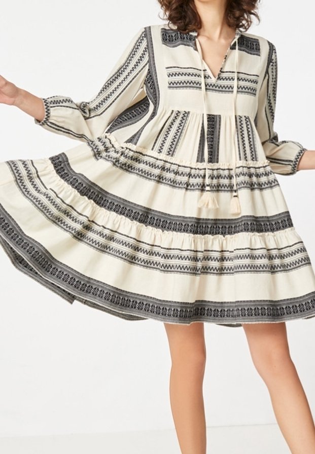 Kvinna som bär en långärmad klänning i krämfärgat och svart mönster med en lager-på-lager kjol, som lyfter fållen för att visa dess flöde och tygdesign.