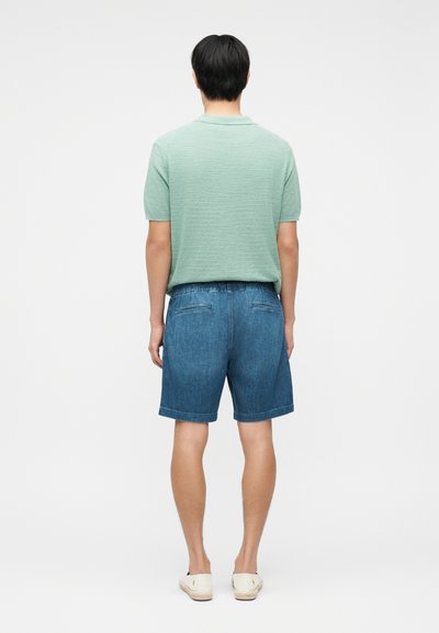 Άνδρας μοντέλο όρθιος με την πλάτη γυρισμένη, φοράει ανοιχτό πράσινο κοντομάνικο πουκάμισο, μπλε τζιν σορτς και λευκά παπούτσια slip-on.