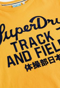 Gele katoenen sweatshirt met gedurfde marineblauwe tekst: "SuperDry TRACK AND FIELD" en extra tekst in het Japans, geribde kraag.