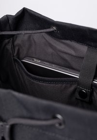 Interno di una borsa nera che mostra un compartimento sottile con un elegante laptop parzialmente visibile. Tessuto morbido e testurizzato con chiusura a coulisse.