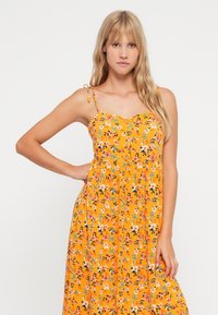 Vestido de verão floral em amarelo brilhante com alças finas, apresentando um decote em forma de onda e pequenos padrões florais em rosa, azul e verde.