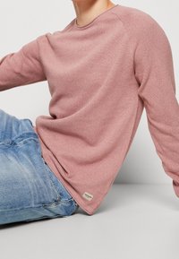 Sweter z długim rękawem, w teksturowanym materiale w kolorze przykurzonego różu. Posiada okrągły dekolt i lekką tkaninę. Połączony z jasnoniebieskimi dżinsami.