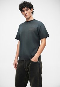 Tricou gri închis din material moale, cu o croială boxy, având un patch discret cu logo pe piept și mâneci scurte, asortat cu blugi negri din denim.