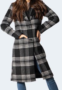 Žena v dlouhém kabátu s černo-bílým kostkovaným vzorem, s knoflíky, s rukama v kapsách, v kombinaci s modrými džínami na světlém pozadí.