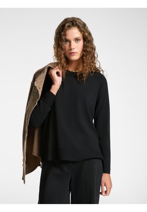 Maglia nera a maniche lunghe con scollatura rotonda, abbinata a un giubbotto color sabbia. Il tessuto sembra morbido con una finitura liscia. Sottolinea una vestibilità rilassata.