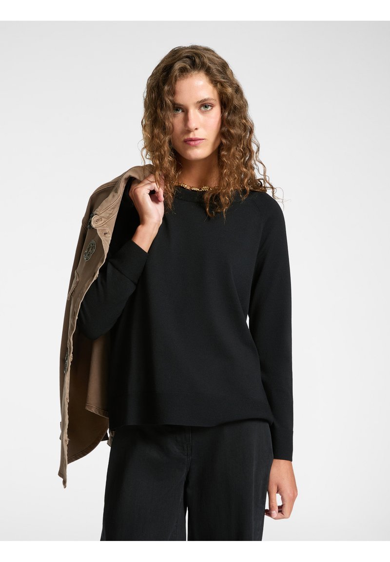 Maglia nera a maniche lunghe con scollatura rotonda, abbinata a un giubbotto color sabbia. Il tessuto sembra morbido con una finitura liscia. Sottolinea una vestibilità rilassata.