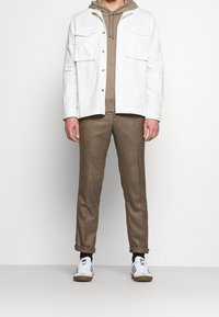 Giacca di jeans bianca sopra un hoodie beige, abbinata a pantaloni marroni e sneakers bianche con dettagli neri. Presenta due tasche sul petto.