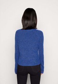 Kobieta z prostymi, ciemnymi włosami, ubrana w dopasowany niebieski sweter z długim rękawem i czarne spodnie, stojąca na tle jednolitego białego tła.