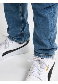 Zapatillas blancas con detalles en negro y un logo dorado. Vaqueros de mezclilla azul claro con un ligero dobladillo en el bajo, que presentan costuras clásicas.