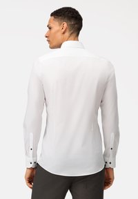 Camicia bianca a maniche lunghe, tessuto liscio, colletto classico, polsini con bottoni e abbottonatura nera, vestibilità su misura, orlo leggermente curvato.