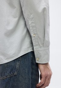 Κοντινό πλάνο στο μπράτσο ενός ατόμου που φοράει ανοιχτό γκρι πουκάμισο με κουμπιά και φαίνεται η τσέπη από σκούρο μπλε τζιν παντελόνι.