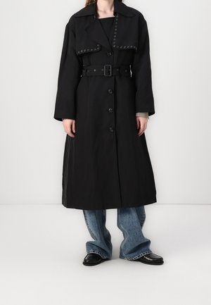 Femme portant un long trench-coat noir ceinturé avec un col clouté, sur un jean bleu et des mocassins noirs, debout sur fond blanc.