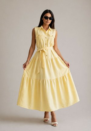 Femme portant une robe maxi jaune sans manches boutonnée avec une ceinture nouée à la taille, des talons blancs ouverts à l'avant, tenant les côtés de la jupe, portant des lunettes de soleil noires.