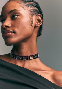 Lila Samt-Choker mit geometrischen Metallakzenten, die Kreise und Diamanten zeigt, getragen von einem Modell mit geflochtenen Haaren und einem schwarzen Off-Shoulder-Oberteil.