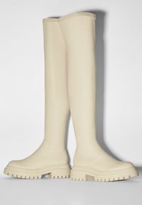 Botas de goma color crema hasta la rodilla con suela gruesa, detalles texturizados y acabado suave. Presenta un diseño minimalista sin herrajes visibles.
