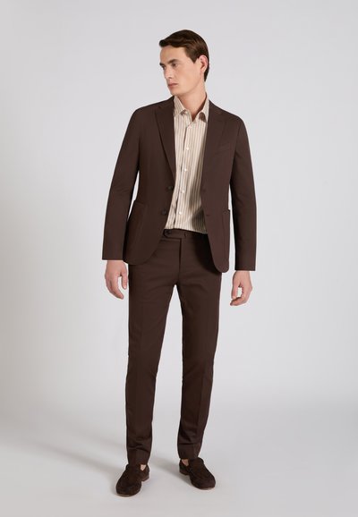 Homme debout portant un costume marron, une chemise beige rayée et des mocassins marron assortis, regardant sur le côté sur un fond uni.