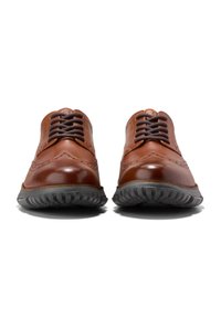 Hnědé kožené společenské boty s brogue detaily, kontrastními tmavými šňůrkami a flexibilní černou gumovou podrážkou. Pohled zepředu ukazuje design.
